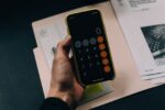 A hand holding a smartphone displaying a calculator app over a paper folder