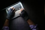 A person wearing fingerless gloves typing on a laptop on a dark surface